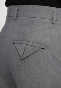 Close-up van grijze broek met een driehoekige klep die gedeeltelijk de achterzak bedekt en zichtbare ceintuurlussen.