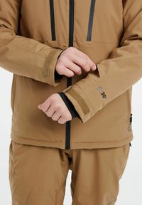 Chaqueta impermeable marrón con acentos negros, cuenta con puños ajustables con Velcro y una calificación de impermeabilidad "20K" mostrada en la manga.
