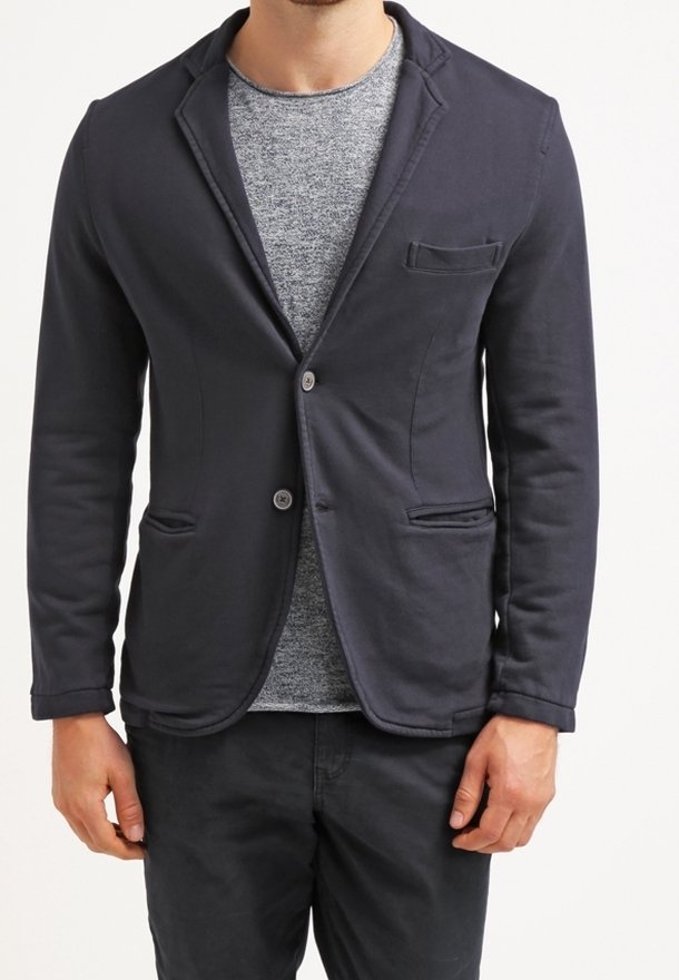 Blazer in misto cotone blu navy con rever a lingua, due tasche frontali e chiusura con un bottone, indossato sopra una camicia grigia a trama.