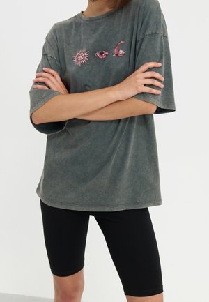 Personne portant un t-shirt gris ample avec des motifs de soleil, œil et lune et un short noir ajusté jusqu'au genou, bras croisés.