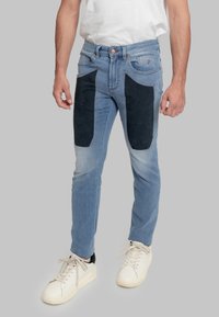 Jeans di denim blu con pannelli in camoscio blu scuro sulla parte anteriore, vestibilità slim, vita a metà altezza e cuciture a contrasto. Scarpe da ginnastica bianche completano il look.