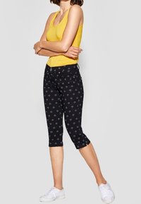 Svart capribyxor med ett vitt geometriskt mönster, gjorda av stretchigt tyg. Stylad med en gul tanktop och vita sneakers.