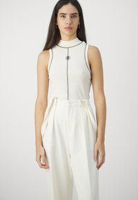 Fitted cream sleeveless top with ribbed texture and contrasting black stitching; high neckline, featuring a logo at the center. White pleated pants.