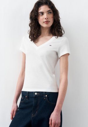 Γυναίκα με σγουρά μαλλιά που φοράει λευκό T-shirt με V-λαιμόκοψη και κοντά μανίκια και σκούρο τζιν ψηλόμεσο, μπροστά σε απλό ανοιχτό φόντο.