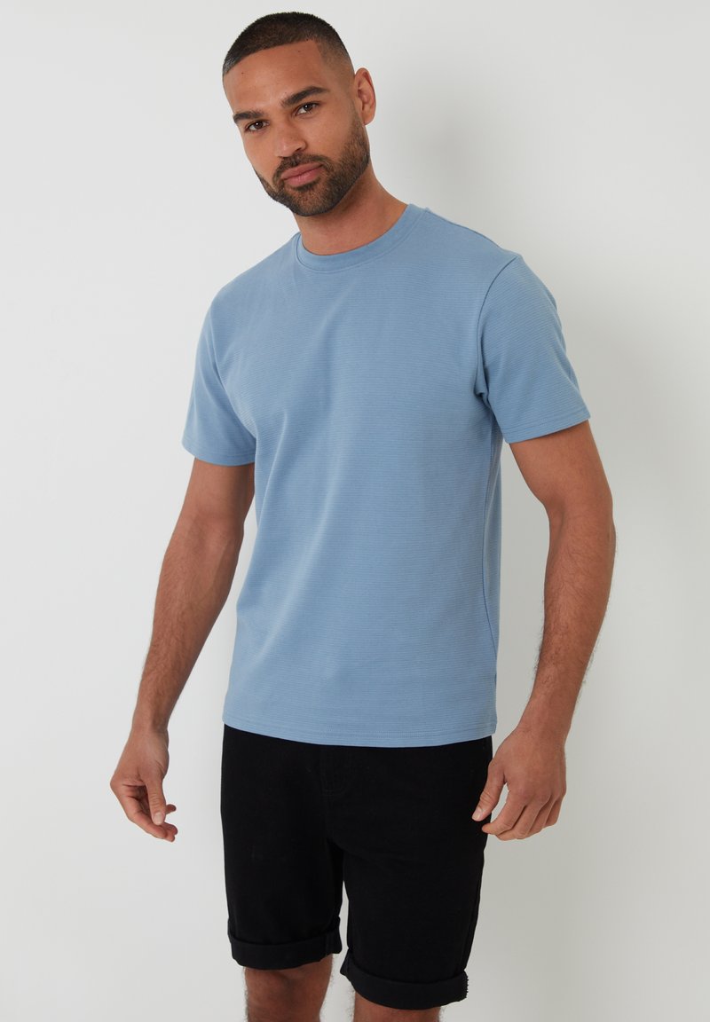 Světle modré tričko s krátkým rukávem a kulatým výstřihem, vyrobené z texturované látky, v kombinaci s černými šortkami s ohrnutím.
