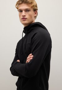 Felpa nera con zip e colletto alto, cordini e una texture morbida. Presenta un design aderente e un aspetto elegante e minimalista.