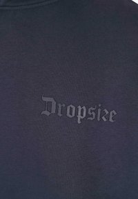 Tmavohnedá látka s nápisom "Dropsize" vytlačeným v gotickom štýle v o niečo svetlejšej šedej farbe blízko hornej časti hrudníka.