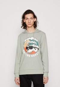 Felpa verde chiaro con cappuccio, caratterizzata da una grafica di un teschio con occhiali da sole, onde e il testo "MAKING WAVES" sotto.