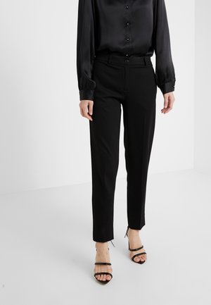 Femme portant un pantalon noir sur mesure, un chemisier en satin noir boutonné et des sandales noires à talons hauts et orteils ouverts, debout sur un sol blanc.