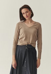 Pull beige à manches longues et col en V, associé à une jupe en denim foncé avec un lien noué, une fermeture boutonnée sur le devant et des détails plissés.