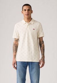 Cremfarbener Polo-Shirt mit einem dezenten Muster, kurzen Ärmeln und einem Kragen. Mit einem kleinen roten Levi's-Logo auf der linken Brust. Getragen mit blauen Jeans.