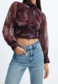 Blouse maroon à motif paisley avec des manches longues transparentes, col haut et taille froncée, associée à un jean taille haute bleu clair.