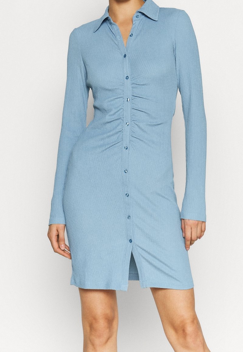 Femme portant une robe ajustée bleu clair à manches longues, boutonnée, avec un col et des détails froncés à l'avant.