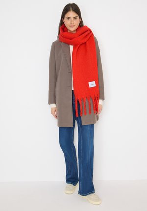 Écharpe en laine rouge avec des bords frangés, portée drapée sur un manteau gris et un pull léger. Présente une petite étiquette avec "CECEL".