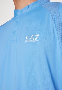 Nära bild av ljusblå sportskjorta med tryckknappar och vit EA7 Emporio Armani-logga på bröstet.