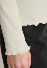 Kremowa prążkowana bluzka z długim rękawem, z falbankowatym wykończeniem na dole i mankietach, zestawiona z czarnym deniem. Skupienie na fakturze i kontrastujących warstwach materiałów.