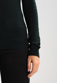 Ciemnozielony sweter robiony na drutach z prążkowanymi mankietami i dolnym brzegiem, posiadający dopasowany krój i miękką fakturę, prezentowany na dłoni i torsie.