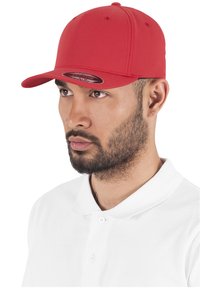 Piros sapka lapos karimával, sima anyagból készült, szellőző lyukakkal rendelkezik, és egy kör alakú logóval van ellátva az elején.