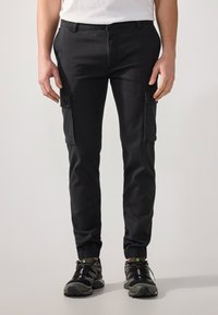 Pantaloni cargo neri con polsini elasticizzati, tasche laterali e una texture liscia. Indossati con scarpe da ginnastica nere con dettagli verdi.