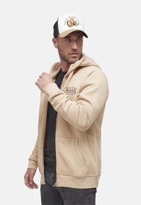 Un sweat à capuche zippé beige avec une poche avant, fabriqué dans un matériau doux, porté par-dessus un t-shirt noir. Accentué d'un logo. Design décontracté. Casquette noire avec un graphique.