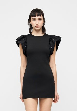 Ung kvinne med skulderlangt mørkt hår som bærer en tettsittende svart kjole med volangerm dekorert med perler mot en ensfarget bakgrunn.