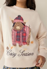Sudadera de color beige claro con un dibujo de una vaca de las tierras altas marrón con un sombrero y bufanda de cuadros rosas, con la inscripción “Cosy Season” en una elegante caligrafía.