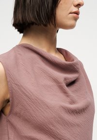 Γυναίκα με κοντά σκούρα μαλλιά που φορά αμάνικο τοπ σε μωβ αποχρώσεις με ντραπέ λαιμόκοψη, μπροστά σε σκέτο φόντο.