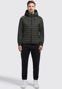 Dunkelgrüne Kapuzen-Pumpsjacke mit gestepptem Vorderteil, Reißverschluss und zwei Seitentaschen. Kombiniert mit schwarzen Jogginghosen und weißen Turnschuhen.