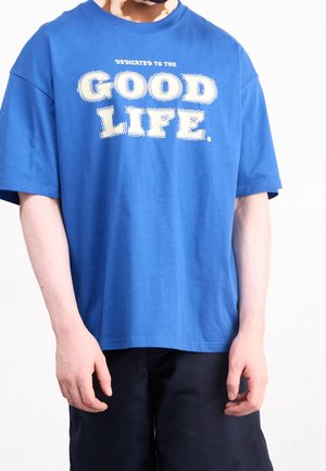 Blue cotton t-shirt with short sleeves and a loose fit. Features white text reading "DEDICATED TO THE GOOD LIFE." on the front.