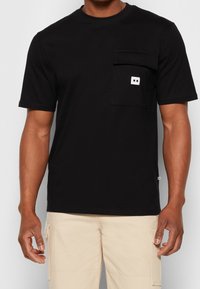 Homme portant une chemise noire à manches courtes avec une poche poitrine et un pantalon cargo beige, visible du cou jusqu'au milieu des cuisses.