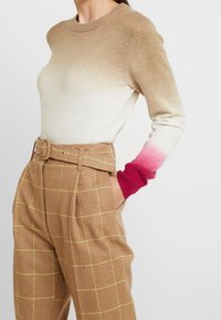 Beige och krämfärgad gradienttröja med rosa detaljer, rundhalsad och ribbade ärmmuddar, tillsammans med bruna rutiga högmidjade byxor med bälte.
