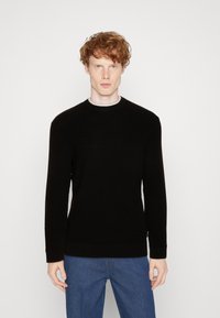 Schwarzer Strickpullover mit rundem Ausschnitt und gerippten Bündchen. Getragen über einem grauen Unterhemd, kombiniert mit blauen Jeans. Einfaches Design.