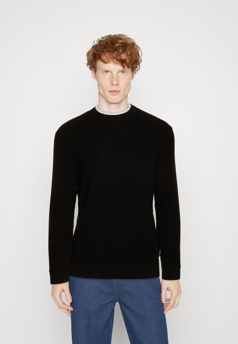 Schwarzer Strickpullover mit rundem Ausschnitt und gerippten Bündchen. Getragen über einem grauen Unterhemd, kombiniert mit blauen Jeans. Einfaches Design.