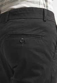 Black pants with a back pocket featuring a button closure and subtle embroidered logo. The fabric appears smooth with a slight shine.
