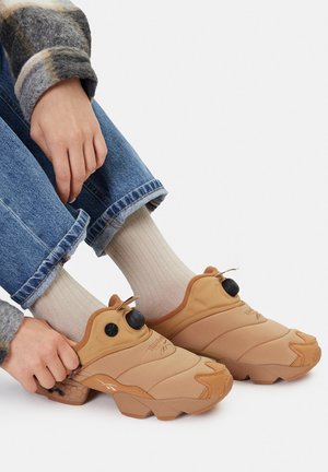 Osoba nosząca jasnobeżowe skarpetki, niebieskie dżinsy oraz poprawiająca jasnobrązowe wsuwane trampki z czarnymi regulowanymi pokrętłami i grubymi podeszwami.