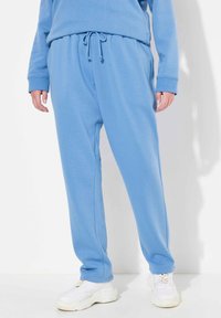 Pantalones deportivos de color azul claro, hechos de una tela suave, con una cinturilla elástica y cordón ajustable, además de bolsillos laterales. Combinados con zapatillas blancas.