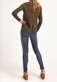 Suéter acanalado verde oliva de manga larga, combinado con jeans de denim oscuro ajustados. Sandalias de tacón cuadrado marrones completan el look.