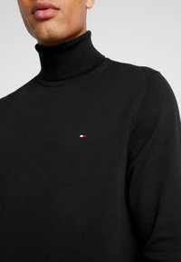 Suéter negro de cuello alto hecho de material de punto, con un pequeño logo que presenta acentos en rojo, blanco y azul en el pecho.
