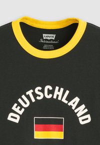 Μαύρο T-shirt με κίτρινο γιακά, που δείχνει λευκό κείμενο "DEUTSCHLAND" και γραφικό της γερμανικής σημαίας κάτω από το κείμενο.
