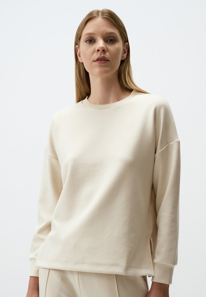 Felpa color crema, in morbido cotone, vestibilità rilassata, scollatura a girocollo, spalle cadenti, spacchetti ai lati dell'orlo, design minimalista.