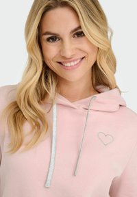 Lys rosa hettegenser laget av myk stoff, med en sølvfarget snøring og en sølvfarget hjertebrodering på venstre brystområde.