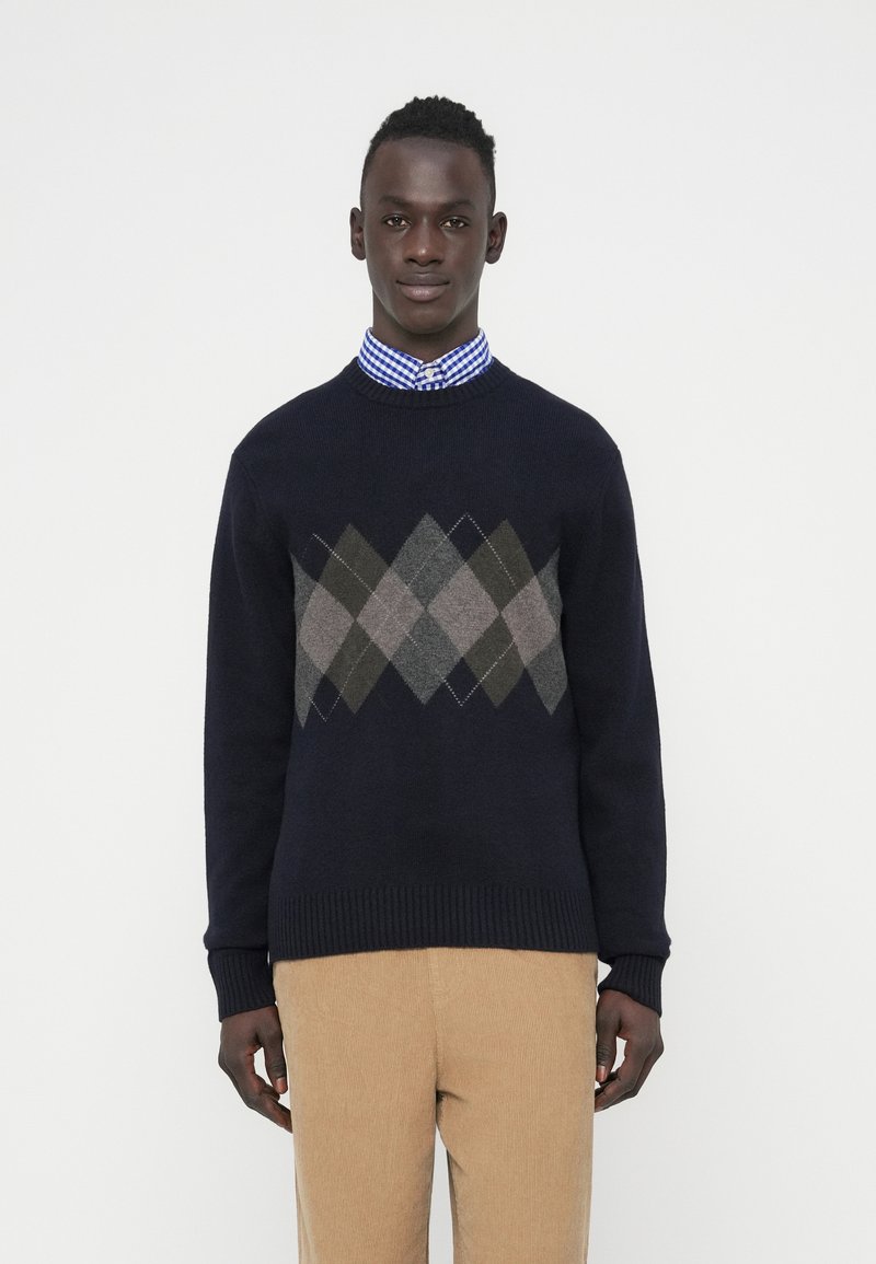 Man wearing a navy blue argyle-patterned sweater over a blue and white checkered shirt, paired with beige corduroy pants, standing against white background.