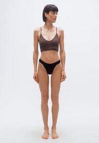 Γυναίκα που στέκεται ξυπόλυτη μπροστά σε απλό φόντο, φοράει καφέ κορυφαίο τοπ και μαύρο ψηλόμεσο εσώρουχο, κοιτάζοντας προς τα δεξιά της.