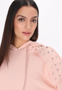 Felpa rosa con cappuccio regolabile, adornata con punte argentate e borchie trasparenti sulle spalle. Tessuto morbido con una vestibilità comoda.
