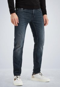 Jean slim-fit bleu foncé en denim, avec un design décoloré, cinq poches, et associés à des baskets blanches.