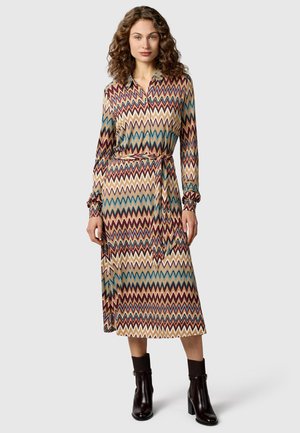 Eine Frau trägt ein langärmliges Kleid mit Zickzackmuster und Gürtel in der Taille sowie schwarze Stiefeletten vor einem schlichten Hintergrund.