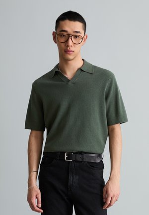 Jeune homme portant de grandes lunettes écaille, une chemise verte à manches courtes avec col, un jean noir et une ceinture noire, debout devant un fond gris.