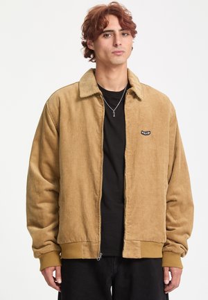 Lysebrun kordjakke med lynlås, ribbet tekstur og et sort logo patch; båret over en sort skjorte. Sorte bukser fuldender outfittet.