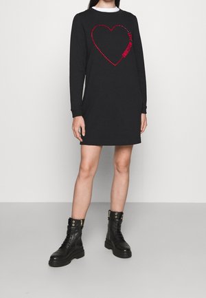 Femme portant une mini-robe noire à manches longues avec un cœur rouge et le texte "Love Moschino", accompagnée de bottines de combat noires à lacets.