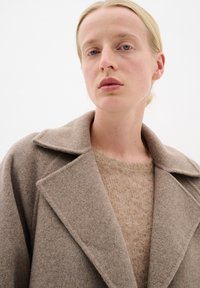Bežna volnena plašča z izrezanim ovratnikom, nošena čez svetlo bež pulover, ki ima mehkobo in minimalističen dizajn. Prikaz od blizu.